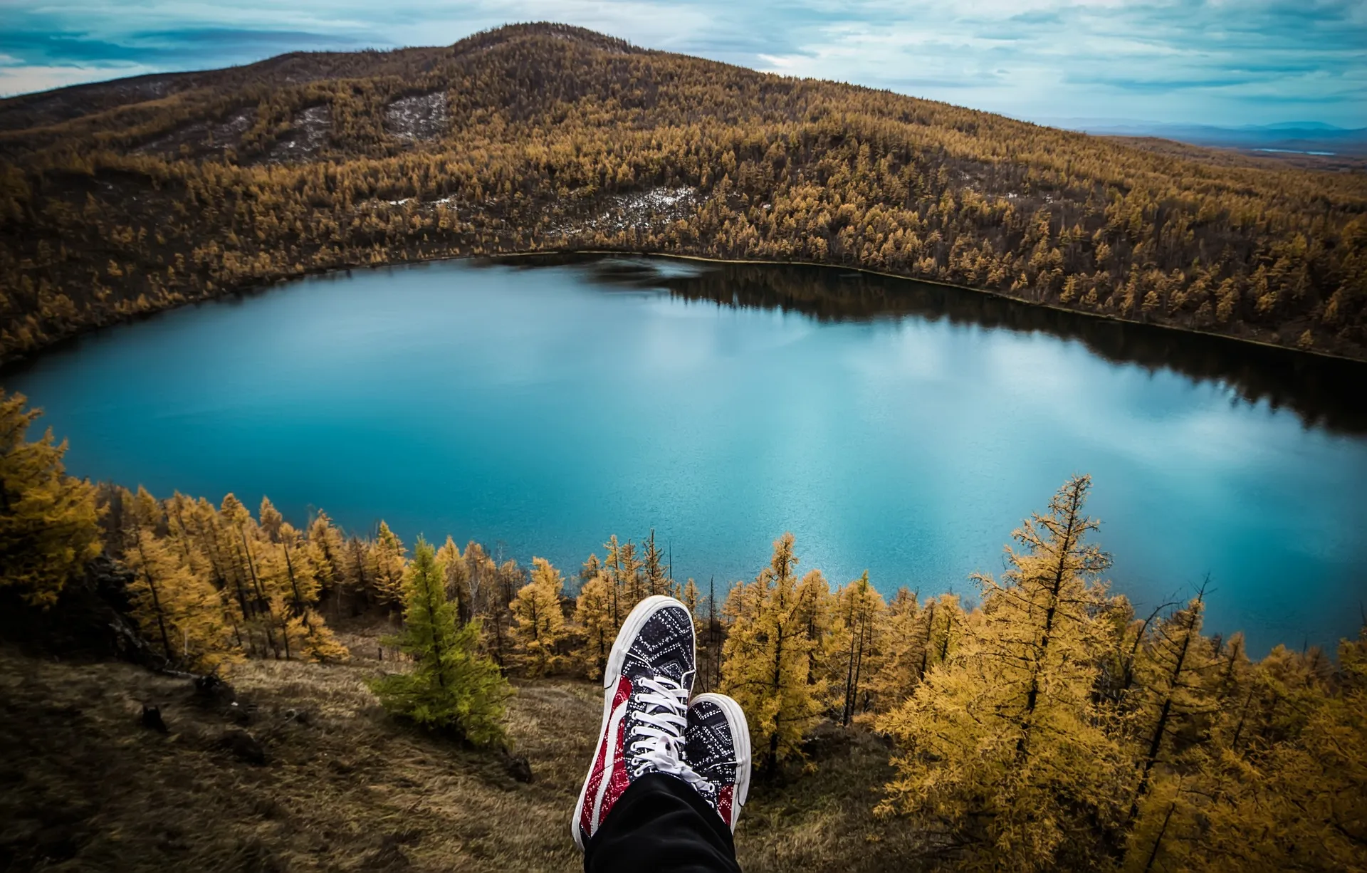 Unos pies con zapatillas asoman sobre un lago rodeado de bosques y montañas.